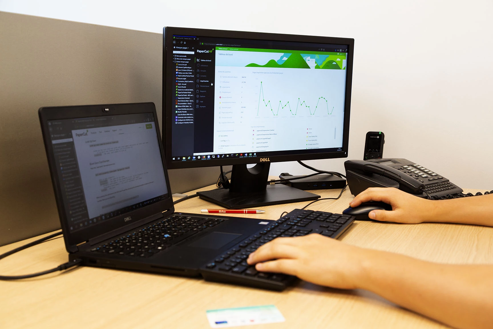 L'image montre un espace de travail avec un ordinateur portable et un moniteur affichant des données, ainsi qu'une personne