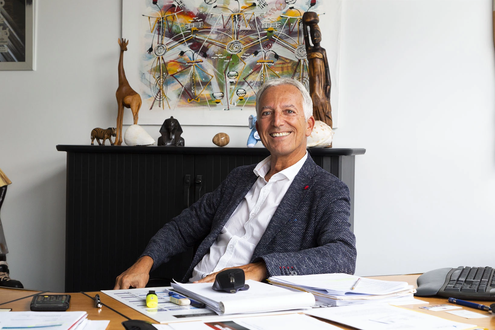 Un homme souriant assis à un bureau avec des papiers et des objets décoratifs en arrière-plan.