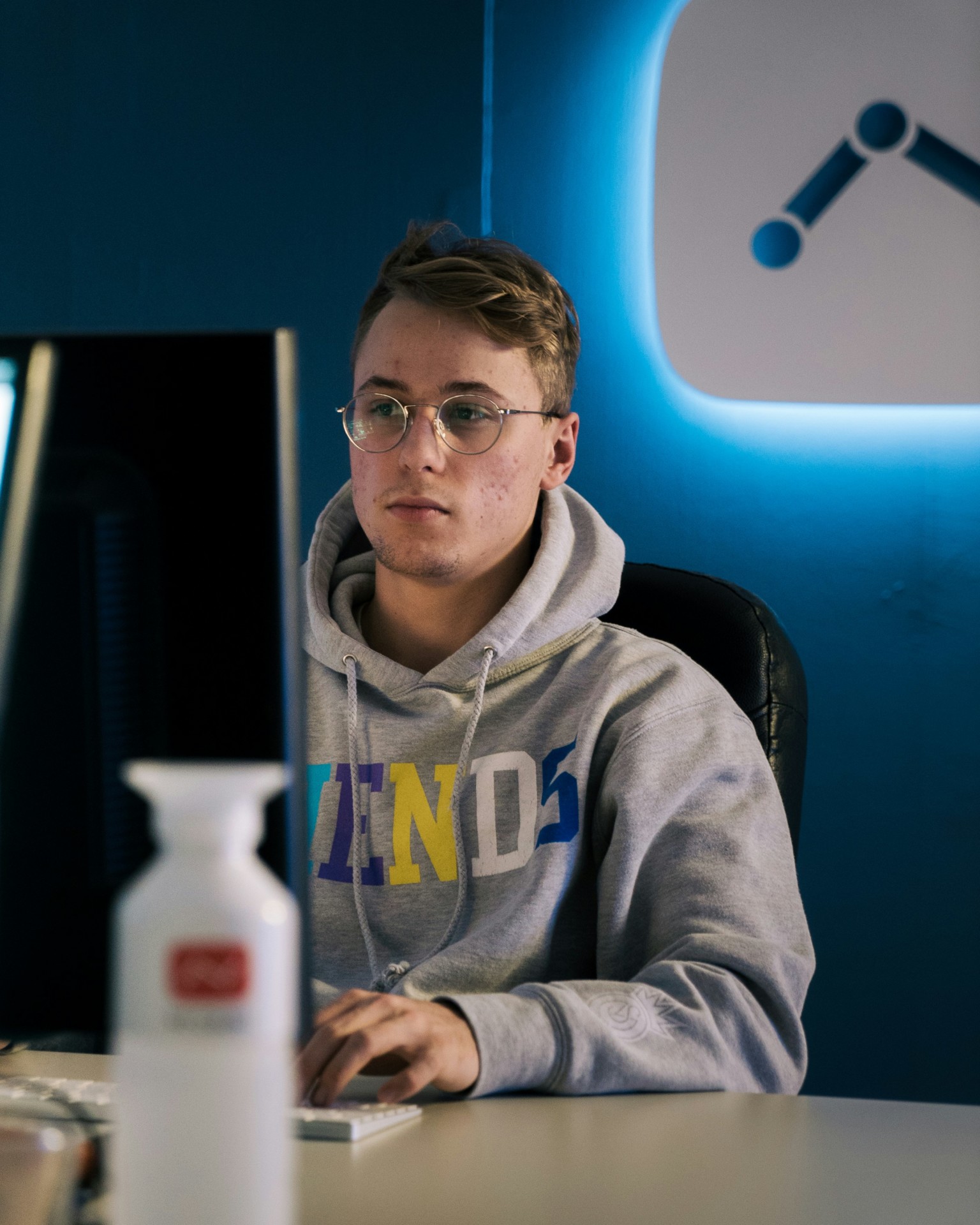 Un jeune homme portant des lunettes et un sweat-shirt gris travaille sur un ordinateur dans un espace moderne avec un mur ble