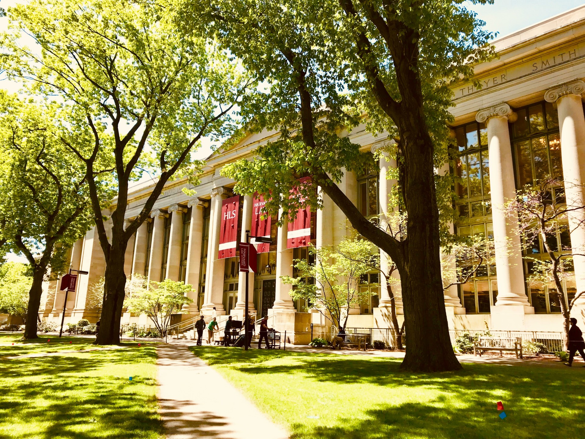 L'image montre un bâtiment universitaire avec des colonnes, entouré d'arbres verts luxuriants et de personnes se promenant 